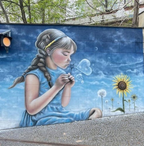 Streetartwall. A beautiful mural of a little girl with soap bubbles was sprayed/painted on a street wall in Odessa. The child with long brown braids in a blue dress is sitting on the ground and blowing a heart-shaped soap bubble into the blue sky. To her right are dandelions and a sunflower. The colors of the Ukrainian flag (blue and yellow) dominate this enchanting little mural.