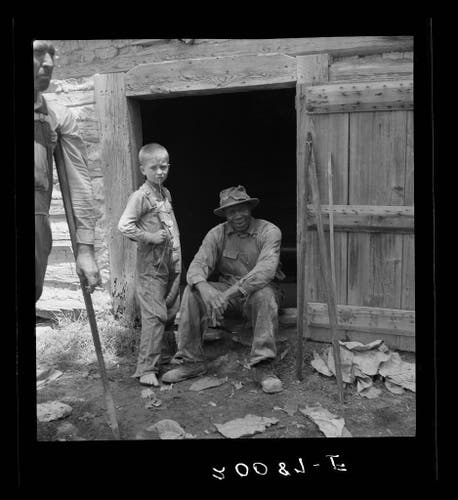  The image depicts a scene of two individuals sitting in front of an old-fashioned wooden structure, which appears to be a barn or storage shed. The person on the left is seated and dressed in what seems to be traditional farm attire, including a hat and overalls, suggesting they might be engaged in agricultural work. On the right, another individual is seated as well, wearing similar clothing that conveys a rustic, rural setting. Both people are looking towards the camera, which gives the impression of a casual or candid photograph.

The background is simple and nondescript, with natural lighting that suggests it might be daytime. There are no visible texts on the image. The quality of the photo has a vintage or aged look to it, with graininess and a slight faded appearance, which could suggest it was taken some time ago.

The context given for this image is "Granville County, North Carolina," suggesting that the setting might be related to agricultural life in that region during a specific period, possibly reflecting traditional lifestyles or activities associated with rural living and farming.