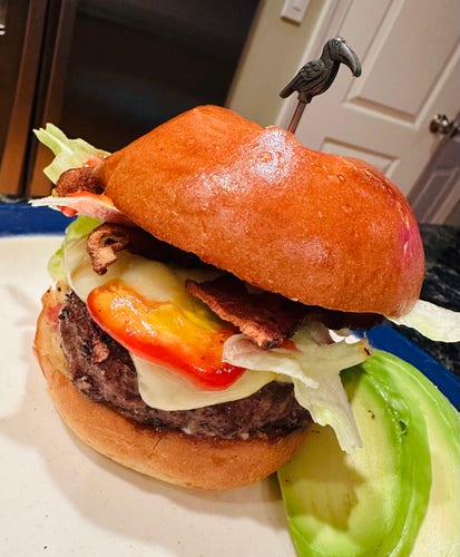 A cute cheeseburger on a round, golden bun with bacon, lettuce, ketchup and mustard. A metal stork toothpick sits on top holding the whole thing together. 