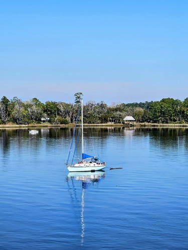Beneath a clear, light blue sky a calm blue river with a tall sailboat anchored in the waters near a lush green shoreline.  The boat's tall mast casts a reflection upon the water below.