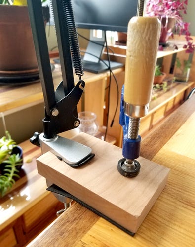 A C-clamp holding a piece of wood onto a table, on which a swivel lamp arm is attached.