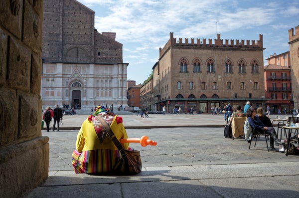 A clown street performer dressed in colorful attire sitting on the ground by a historical building's wall with a toy balloon gun, overlooking a spacious square where people walk and sit at outdoor cafe tables.
