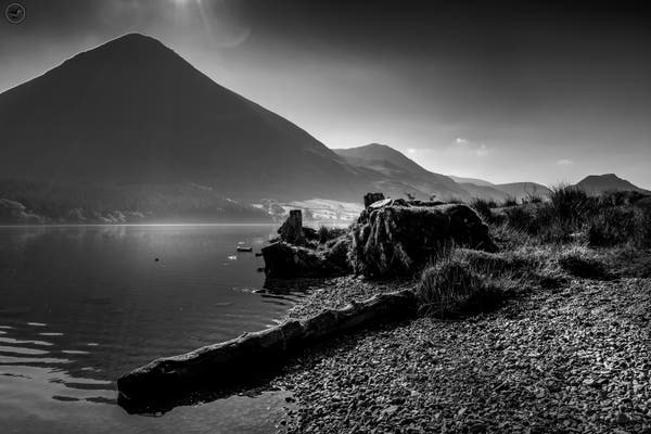 Monochrome shot over lake, with fallen log and stumps in foreground, mountains in background and sun halo peaking over mountain top