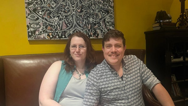 Two people sitting on a brown leather sofa in front of a yellow wall with some kind of surrealist clockwork gear painting. On the left is Dani, with her auburn hair hanging down around her shoulders. She has a slight smirk. To her right is me, her husband Sean. I am flashing a toothy smile.