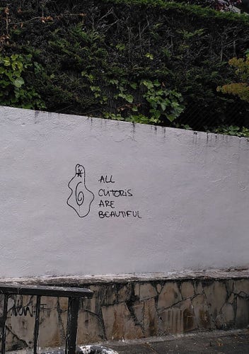 White wall of an underpass. On it in black marker pen, "all clitoris are beautiful" next to a (fairly abstract) sketch.
