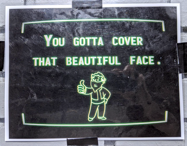 Photo: a sign taped to a wall depicting an image similar to a Fallout Pip Boy screen, computer green on a black background. The image is of VaultTec Corporation's mascot Vault Boy in a mask giving a "thumbs up" with the text reading: YOU GOTTA COVER THAT BEAUTIFUL FACE.
