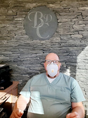 My denturist, Dr. Ken Roberts, at Burnside Dentures Clinic. He's wearing green surgical scrubs & a white N95 mask. He is sitting at reception desk. His company logo is on the wall behind him.