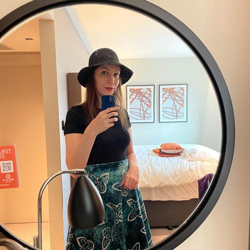 A photo of me, a 40-year-old white woman with blue eyes and long brown hair, wearing a black crew-neck top, a green batik skirt with butterflies, and a black sun hat
