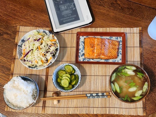 A Japanese meal on small white and blue enamel plates and bowls. There is a slice of salmon, a cabbage salad, miso soup, rice and pickled cukes. The chopsticks rest on a wooden mushroom and my kobo is at the top showing the cover of Welcome to the Hyunam-Dong Bookshop.