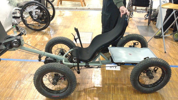 A pale green cargo recumbent quadcycle with a bottom/front bracket engine and fat, knobby tires. The cargo bay is seems a Eurobox bottom.