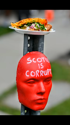 A photo of a red mannequin face with the text "SCOTUS IS CORRUPT" written on the forehead. It is attached to a metal bar, on top of which is a plate with some tacos.