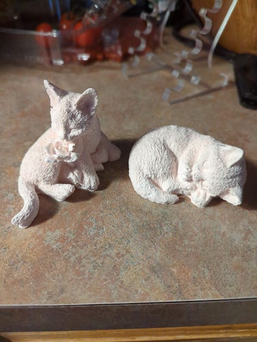 Two small, pinkish plaster casts of kittens. One is lying on its side and sniffing a rose. The other, to the right, is curled up and sleeping.