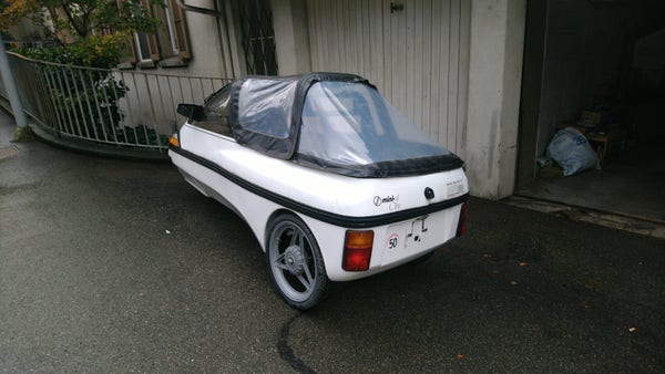 A white three-wheeler electric vehicle from the early 90s with a black "cabriolet" canopy with large transparent parts; standing in front of a garage, view from left back.
