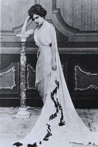  The image appears to be an old photograph featuring a woman posing in formal attire. She is wearing a long, elegant dress with intricate details and what seems to be a sash or shawl draped over one shoulder. Her hair is styled up, and she has a poised posture, resting her hand on a column or pillar behind her. The background suggests an indoor setting, possibly an interior space within a building, indicated by the architectural features visible in the shot. The image is monochrome, which adds to its vintage feel.