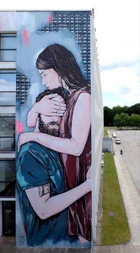 Streetartwall. The mural of a pair of lovers was sprayed/painted on the narrow part of an exterior wall of a four-storey modern building. The mural is designed like a watercolor drawing and in light shades. The background is light blue with a gray grid pattern. It shows a young, dark-haired woman in a red dress embracing a dark-haired man with a full beard in a blue shirt. She has closed her eyes, hugs him to her chest and covers his eyes with one hand. He puts an arm around her. (A strip of grass and a road can be seen next to the mural). Not in the photo is another fantastic large mural on the side of the same house.
Info: The Austrian and French street artists Jana & Js have been painting together since 2006. The artworks are mainly based on their personal photographic work. Inspired by the place where they create their works, they focus on nostalgia and melancholy.