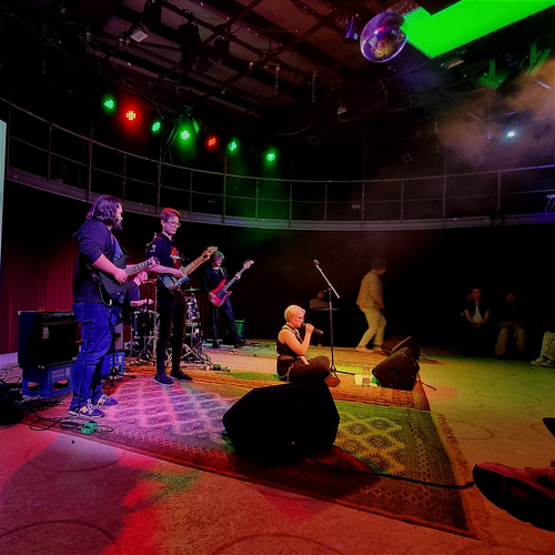 Die Band Spezi im Glas auf der Bühne während des Auftritts. Ria (Gesang) sitzt auf dem Boden in der Mitte und singt. Scheinwerferlicht und Nebel durchsetzen das Bild.