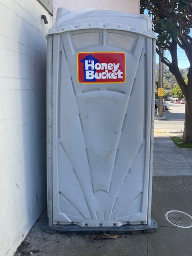 Photo of a grey porta-potty with a large “Honey Bucket” logo on it.