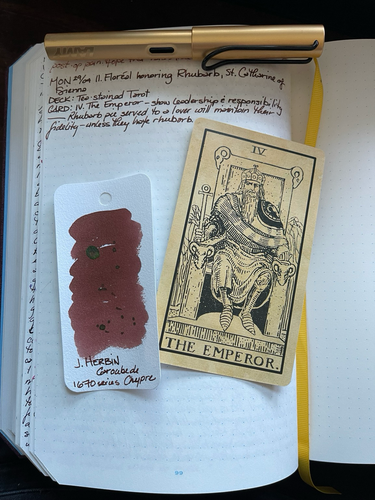 Open notebook with handwritten text from the post with additional text: “Rhubarb pie served to a lover will maintain their fidelity [Cunningham] — unless they hate rhubarb.” Above the text is a gold toned pen with a black clip. Below the text is a swatch card showing a reddish brown ink with green sheen and gold shimmer and the Emperor tarot card with the king sat on his ram’s head throne holding an ankh.