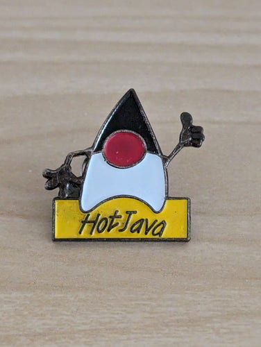 Closeup view of a metal lapel pin on a light wooden surface. The pin has the shape of a cartoon figure with a body and head shaped as a whole like a Starfleet logo, black head and hands, and white body. The left hand of the figure is raised doing the thumb up sign. Below the figure is a plate with the label HotJava.