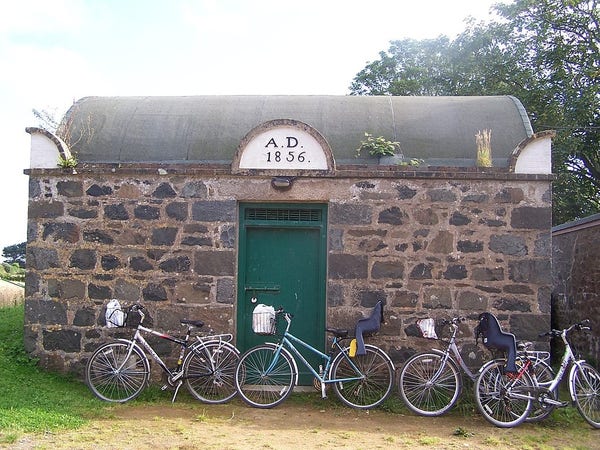 This is a photo from the fiefdom of Sark.

It shows an old building made of large stones with a dome-shaped roof. It has a dark green door. Over the door is a sign, in white, saying "A.D. 1856".

In front of the building are three bicycles.

It is the prison of Sark.