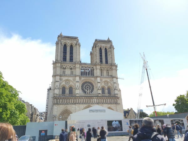 Photo of the Notre Dame cathedral on a bright sunny day. There's a construction site around it, with a big crane on one side.