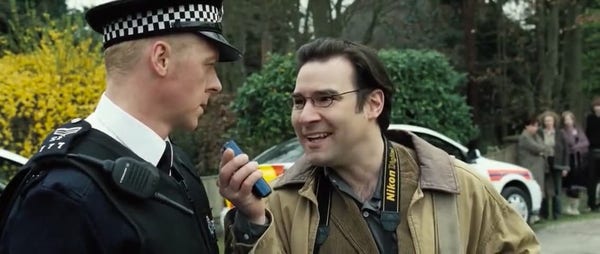 A screenshot from Hot Fuzz. Tim Messenger is holding a dictaphone in front of Nicolas Angel. In the movie he’s asking what’s your perfect Sunday. 