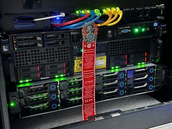 A rack of servers with a router attached. There is a warhammer 40k seal of the The Machine God, also known as the Omnissiah or Deus Mechanicu on the servers to bless them against technical problems.