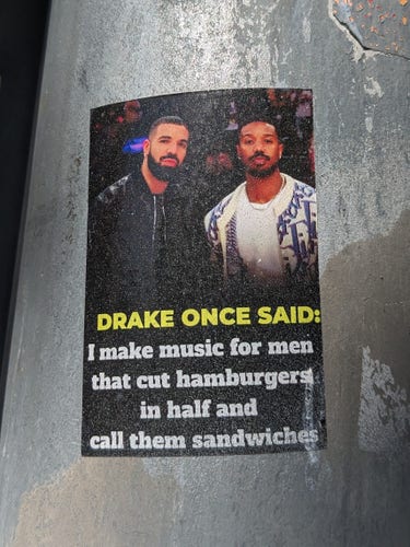 A picture of a sticker on a street pole. It's a photograph of the rapper Drake and someone I don't recognize. There is a caption that reads "Drake once said: I make music for men that cut hamburgers in half and call them sandwiches"