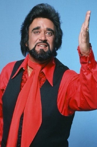 Photo of Wolfman Jack, classic radio era DJ, actor, all around celebrity. He has blow dried styled hair medium length up above his head. Photo appears to be from the 70s so he is wearing a ruffled red silk shirt with a red scarf tied around his neck and a black vest. He also has a beard and mustache, his hair reminded people of the Wolfman from classic horror films. 