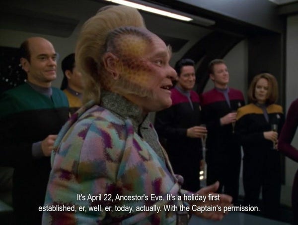An alien with light skin with purple patterns on it, wearing a very colorful suit, is standing with a group of Starfleet officers. He's saying, “It's April 22, Ancestor's Eve. It's a holiday first established, er, well, er, today, actually. With the Captain's permission.”
