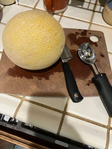 Muskmelon (cantaloupe, probably) next to sharp knife next to a shiny metal scoop atop a cutting board. 