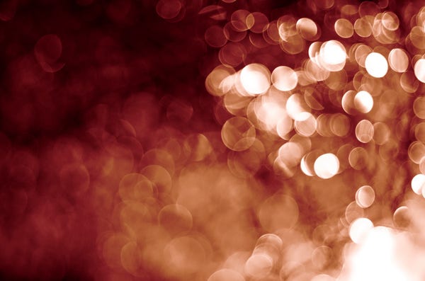 Abstract bokeh lights with a soft focus, predominantly in reddish and sepia tones. The bottom right corner is the brightest, while the upper left is dimmer. It looks like sparks flying up from a fire and disappearing into the night.