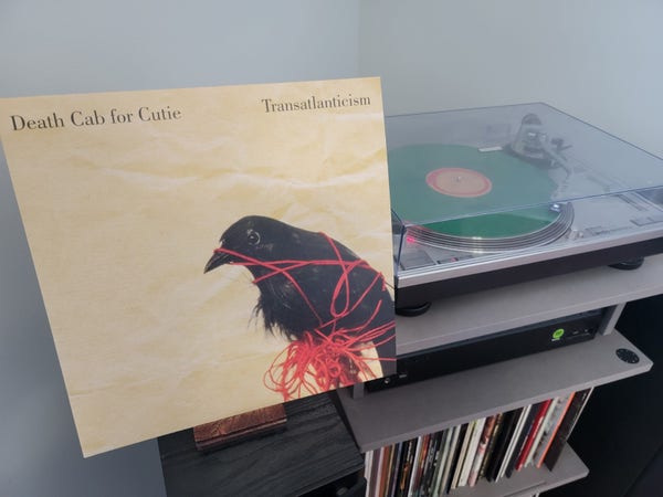 Cover art showing a black bird tied in red yarn over a yellow background. Vinyl in background is green