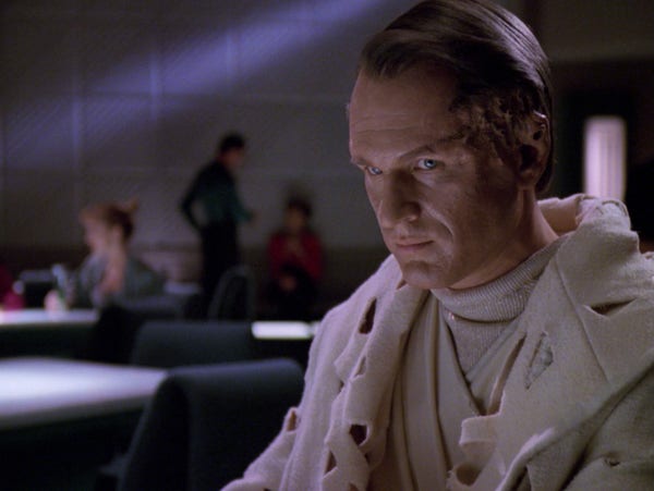 A robed male alien sits in 10 Forward and stares directly into the screen at the TV viewing audience.