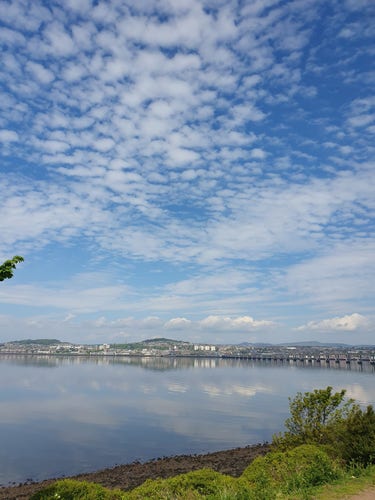 A view towards Dundee, looking over the River Tay. The river has a gentle ripple as the sky comes in, the reflection of the blue sky with white wispy clouds, and the reflection of Dundee can be seen in the river.