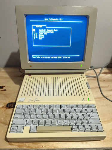 A photo of an Apple //c computer with monitor sitting on a plywood workbench