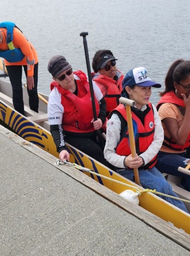 Me in second to last right side of dragon boat, holding paddle. 3 paddlers are around me in other seats. Steering safety person is standing behind us. Boat is still clipped to dock.