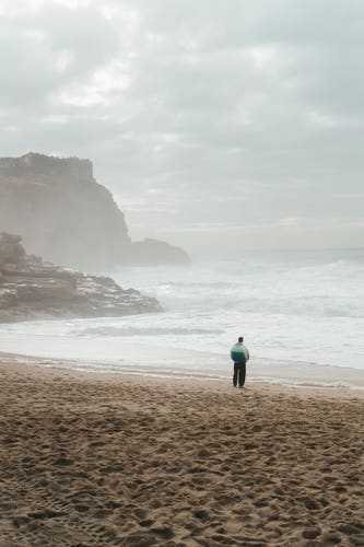 A beach with lots of mist, a man stands on the right side of the photo. In the background is an overcast sky and cliffs.