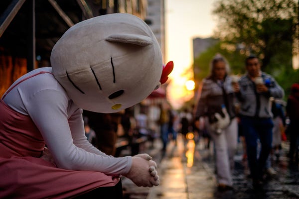 Person in a Hello Kitty costume with a giant fuzzy head and pink dress sits with their head down and hands clasped in front of them on the street. The sun is setting in the background and people are walking by without paying attention to Hello Kitty.