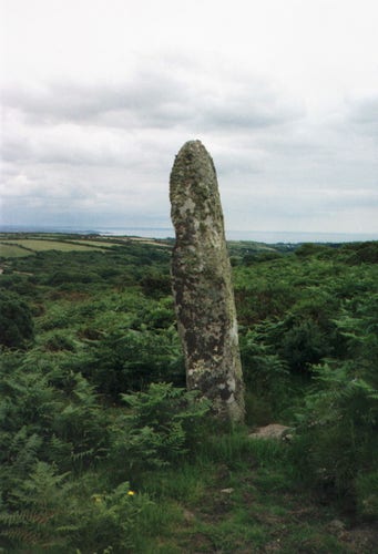 Carfury Menhir in Madron parish. This prehistoric standing stone is about 3m or nearly 10ft tall and is surrounded by ferns and lush dark gree vegetation. Another menhir is close by, but lies prostrate. Mount's Bay can be seen in the distance.