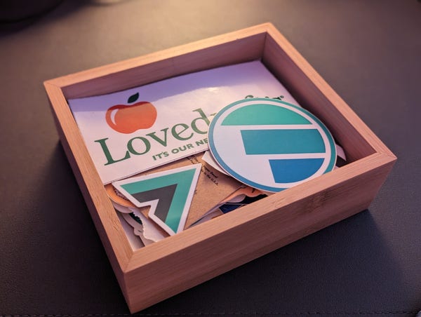 Small, wooden box filled with stickers. Visible on top are logo stickers for Vue and Polypane, and a sticker for a local bar called Lovedraft's, which parodies Applebee's with the tagline: "It's our neighborhood now"