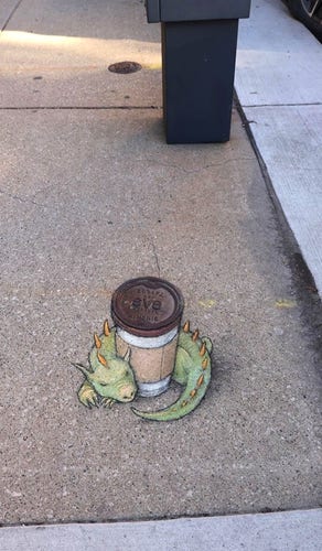 Streetart. A green dragon with a "coffee to go" cup was painted with chalk on a gray sidewalk. The cup is actually a metal gas valve cover in the street, under which a chalk-painted cup has been added. The green dragon with yellow spikes wraps itself around it and keeps it warm.
Title: "Griselda can keep your coffee warm indefinitely, but getting it back will cost you a cruller."