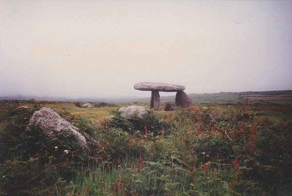 Colour photo of Lanyon Quoit in Cornwall, taken in July 1993, showing the stones at the east end of the mound and with the portal tomb or dolmen at the other end. The mound is surrounded by green ferns and small flowers and the moors surrounding are covered in fog.