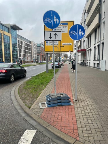 Fahrradweg in Ludwigshafen Rheinstraße. Das Schild versperrt den Radweg...

Automatisch generiert:
Street view with traffic signs indicating bike and pedestrian paths, directional arrows, and parking information. Modern buildings line the road, a pedestrian walks by, and cars pass in the background. Pallets and a cover on the ground suggest maintenance work.