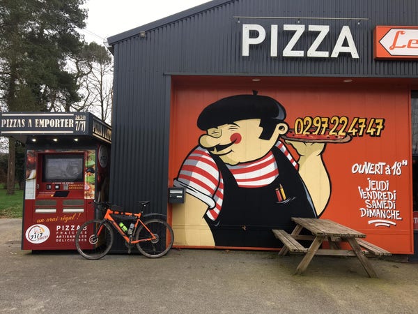A bright orange bike leaning against the wall between a pizza restaurant and the pizza robot -- a device that provides a range of pre-made pizzas when the full-service restaurant is closed.