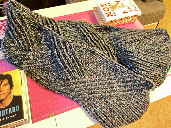 A knitted scarf on a tabletop. The yarn is a variegated blue/purple/grey. The stitching is a zigzagging cable 