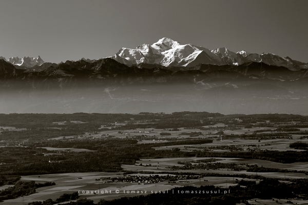 Black and white photograph showing a panoramic view of the Mont Blanc massif. In the foreground are fields, woods and buildings, and just below the massif is a light mist over Lake Leman, which passes smoothly and ends just below the summit of Mont Blanc.