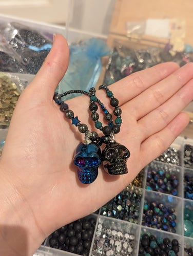 photo go hand holding 2 handmade necklaces with black and skull metallic pendants and beaded necklaces with hematite styled, green and blue star beads