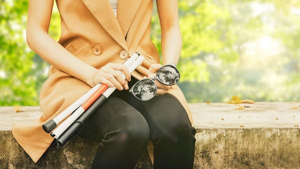 A young woman sits on a stone bench holding sunglasses and a folded white cane in her lap.