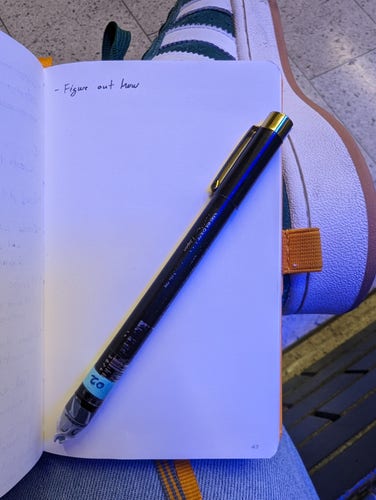 A notebook lies open with a black.pen lying across it. The page is blank save for a small note at the top  which says "figure out how".